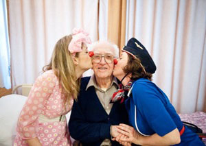two women with glued clown noses kiss an elderly man on the cheeks